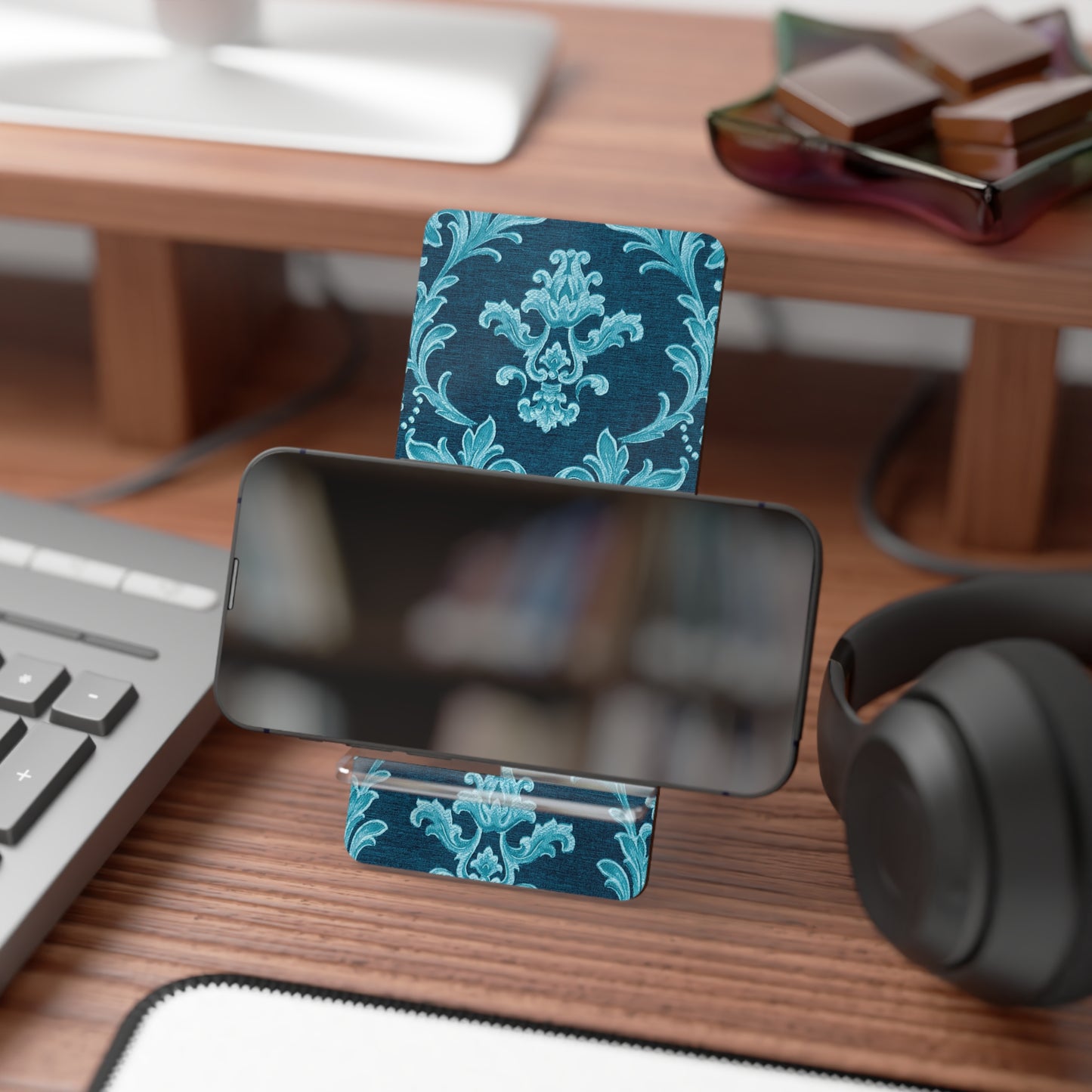 Phone stand with blue floral design on a desk with keyboard, mouse, and headphones.