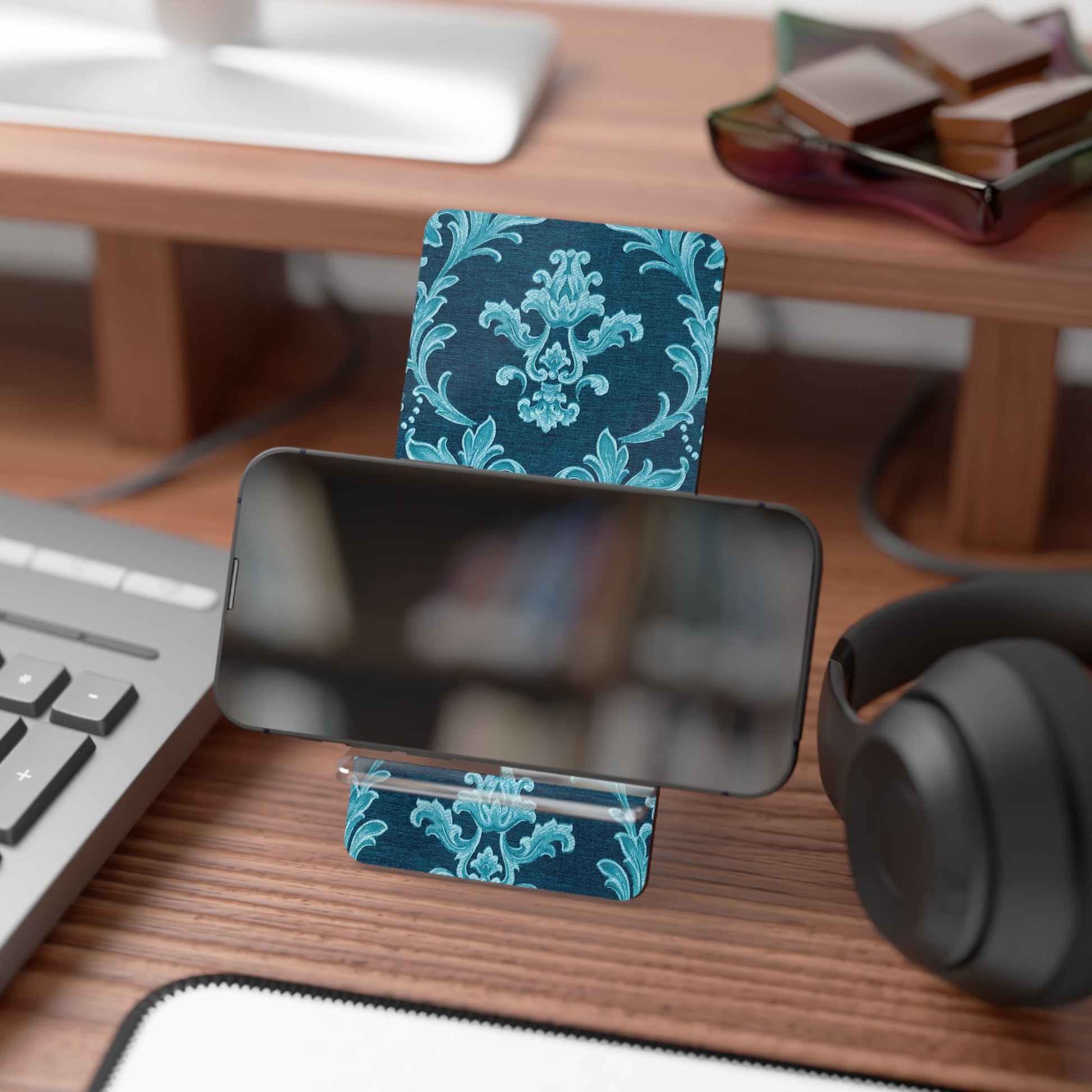 Phone stand with blue floral design on a desk with keyboard, mouse, and headphones.