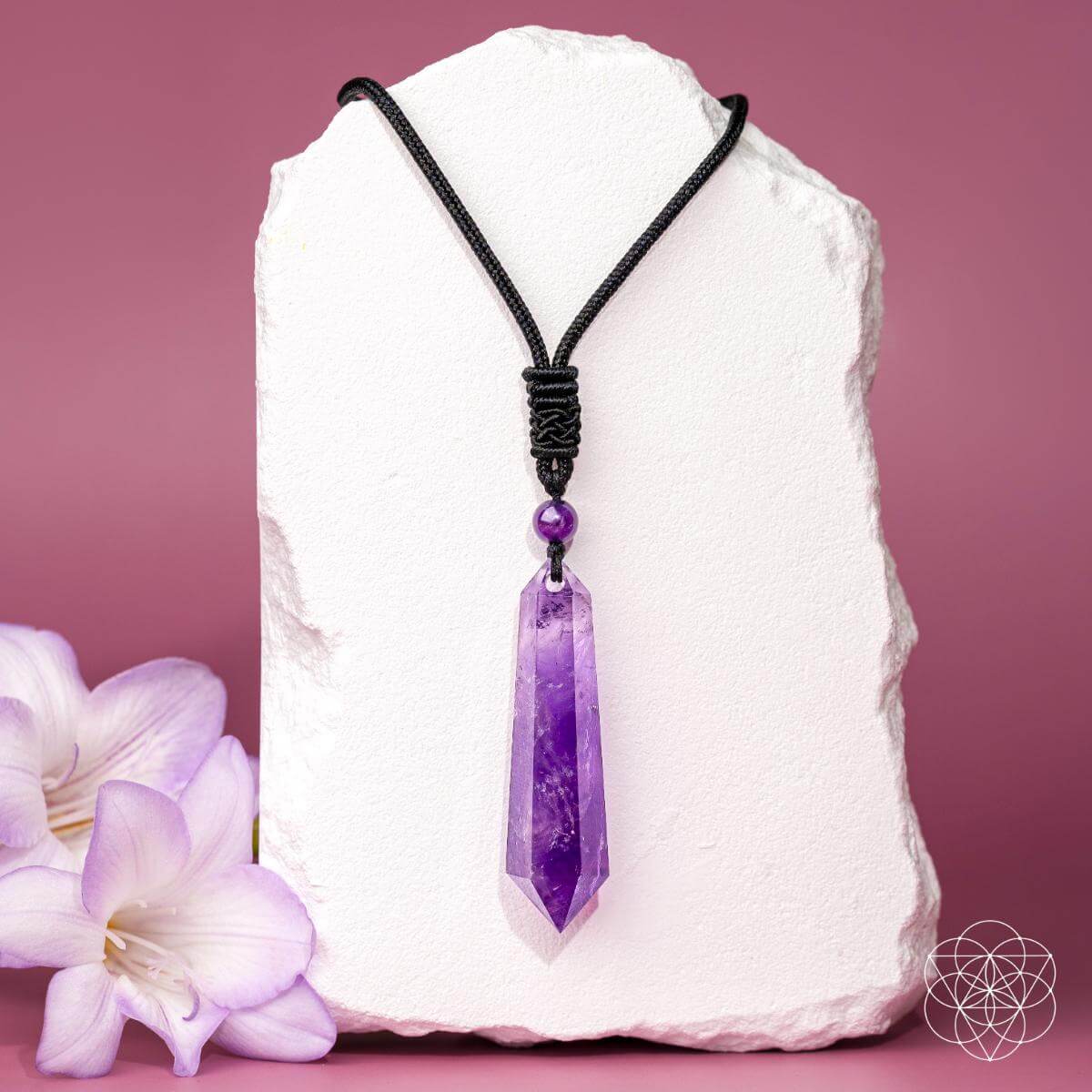 Purple crystal pendant on a black cord with a white stone and purple flowers on a pink background